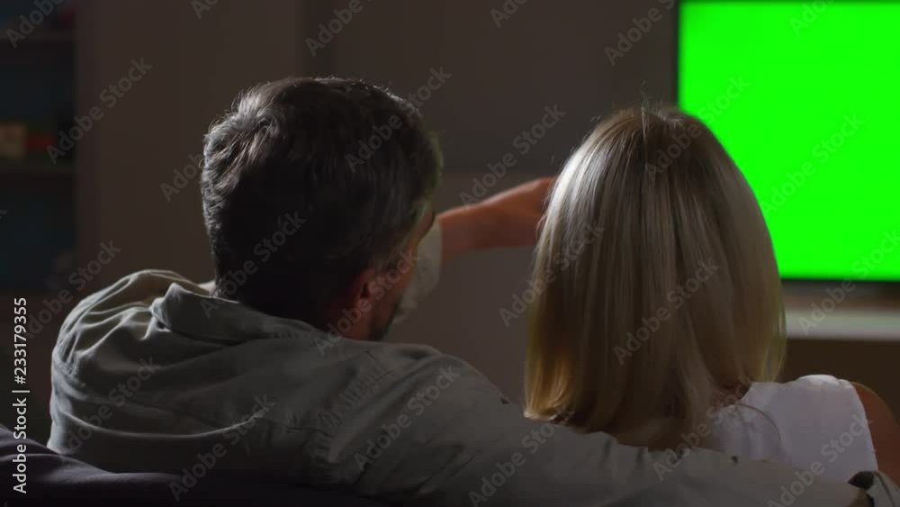 Rear view of husband and wife pointing at green TV screen and discussing something while sitting together on sofa at home and watching movie