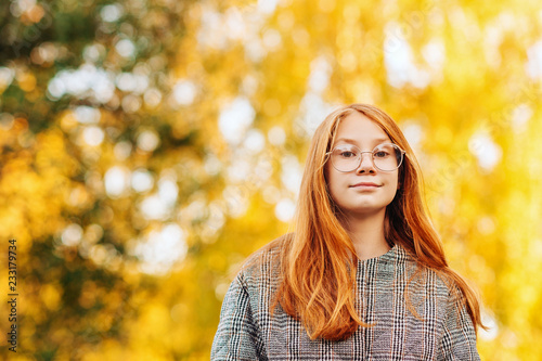 Outdoor close up portrait of beautiful red-haired preteen girl wearing check coat and eyeglasses © annanahabed