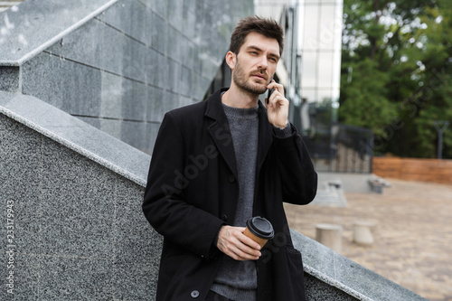 Handsome young business man sitting outdoors talking by mobile phone drinking coffee. © Drobot Dean