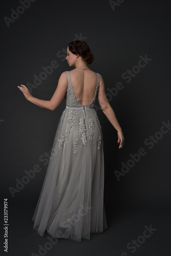 full length portrait of brunette girl wearing beautiful long gown  standing pose with background to the camera on grey studio background.