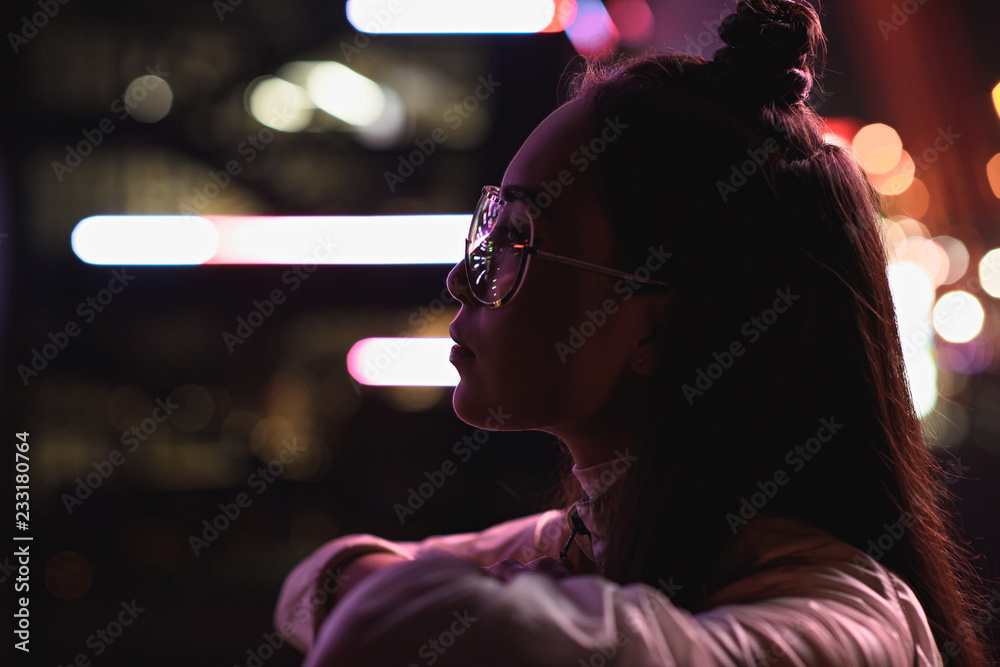 Obraz premium side view of stylish asian girl in glasses looking away on street with neon light in evening, city of future concept