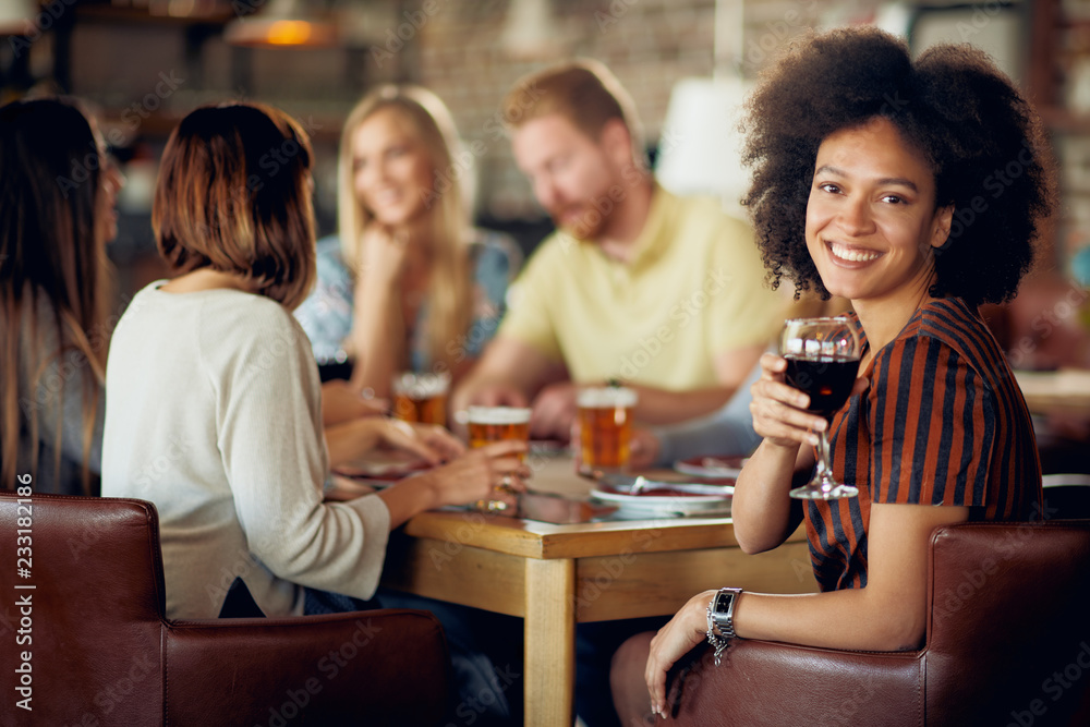 Woman looking at camera and holding glass of wine while sitting in restaurant. In background friends drinking and chatting.