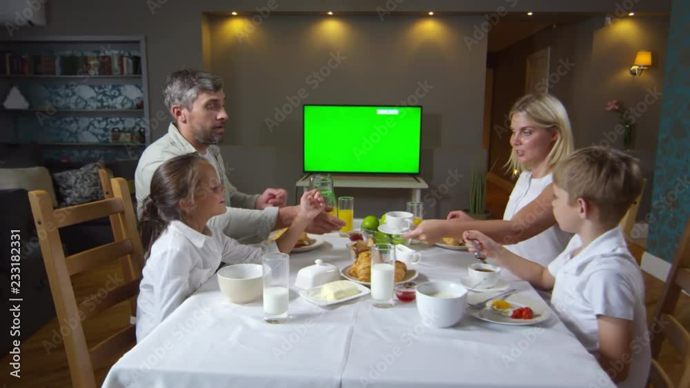 Panning shot of beautiful parents talking with little daughter and son while having breakfast together and watching TV with chroma key green screen Panning shot of beautiful parents talking with little daughter and son while having breakfast together and watching TV with chroma key green screen