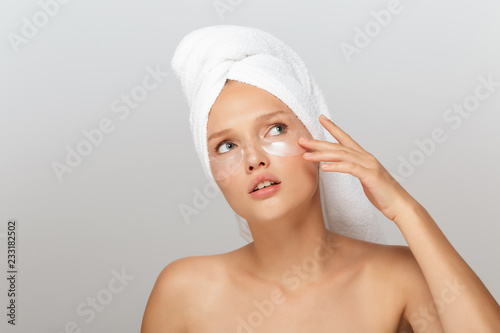 Portrait of young woman with white towel on head without makeup with transparent patches under eyes thoughtfully looking up over gray background isolated