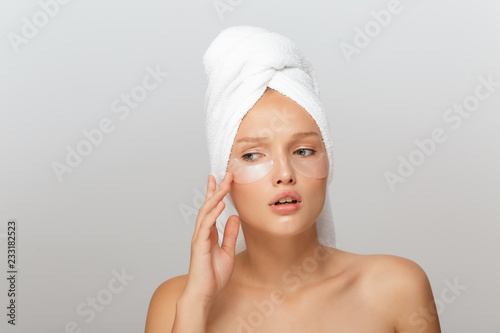 Portrait of young upset lady with white towel on head without makeup with transparent patches under eyes thoughtfully looking aside over gray background isolated
