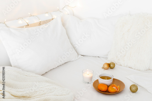 White cozy bedroom with holiday decoration. Bed with white bedding set, pillows, wooden plate, cup of tea, tangerines, christmas balls, candle and garland lights. Home christmas decor.