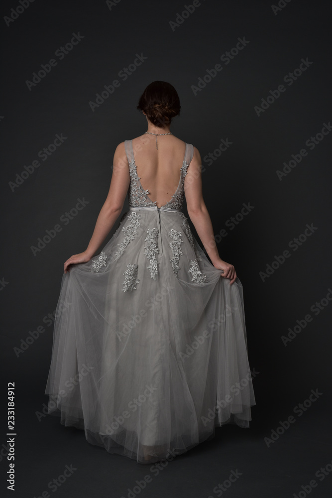 full length portrait of brunette girl wearing beautiful long gown, standing pose with background to the camera on grey studio background.