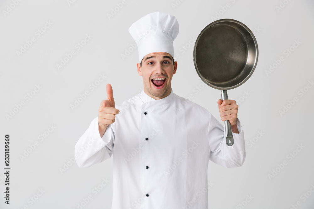 Emotional young man chef indoors isolated over white wall background holding frying pan. Emotional young man chef indoors isolated over white wall background holding frying pan.