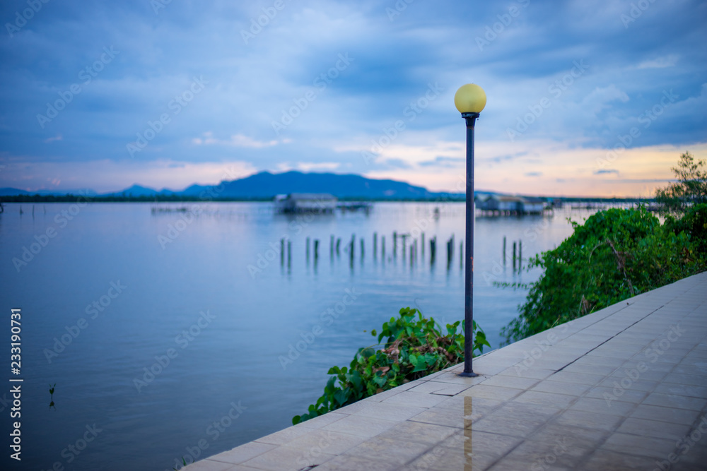 Old Yellow street light post on the walkway near the lake with blurred view of the fisherman house mountain and cloudy sky.