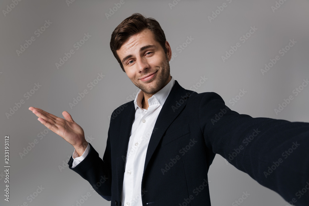 Image of successful man 30s in business suit gesturing hand aside while taking selfie photo, isolated over gray background Image of successful man 30s in business suit gesturing hand aside while taking selfie photo, isolated over gray background