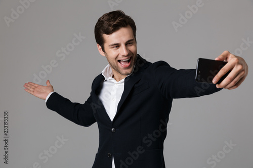 Image of joyous businessman 30s in formal suit holding copyspace on palm while taking selfie photo with mobile phone, isolated over gray background