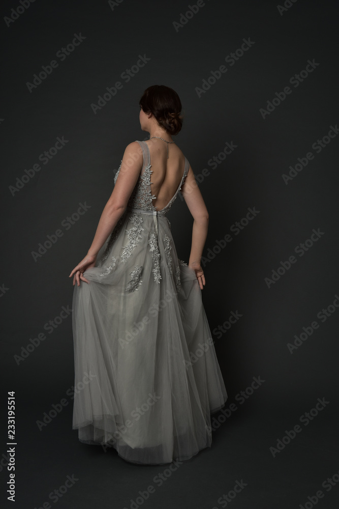 full length portrait of brunette girl wearing beautiful long gown, standing pose with background to the camera on grey studio background.