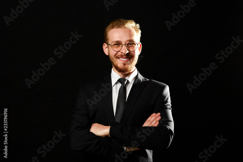Close up portrait of successful man with beard in glasses keeping arms crossed and smiling on black background. Startup and business concept.