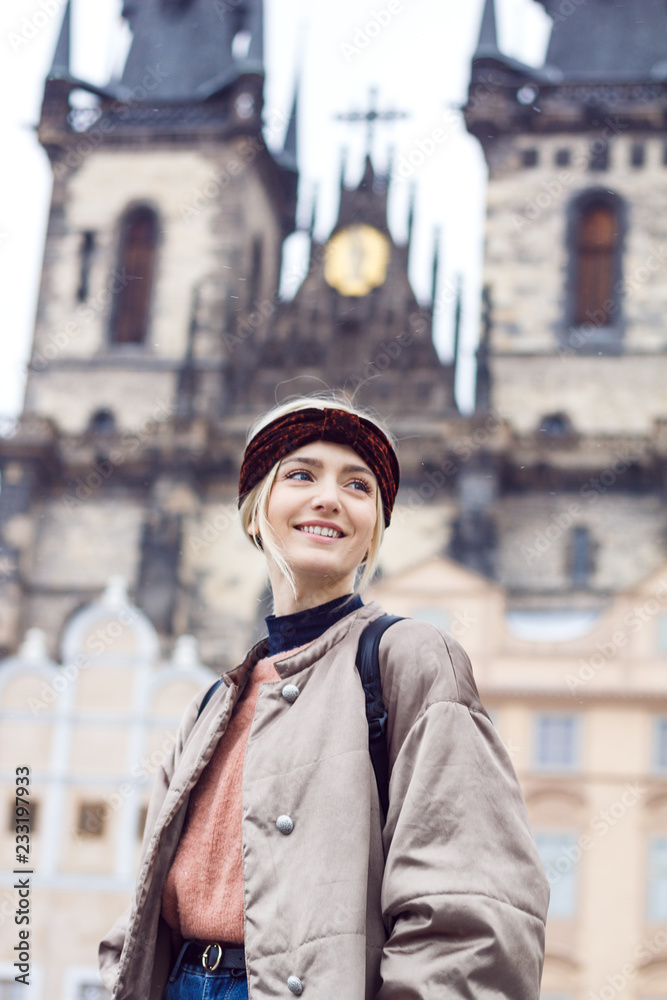 Portrait of a beautiful blonde woman in a cold winter in the historic district. She's wearing a warm winter jacket, pink sweater, jeans, backpack and ears are covered by headband. Portrait of a beautiful blonde woman in a cold winter in the historic district. She's wearing a warm winter jacket, pink sweater, jeans, backpack and ears are covered by headband.