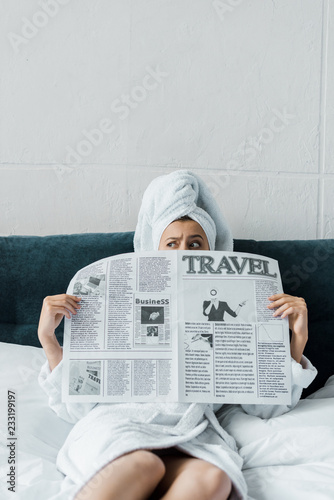 confused girl hiding behind travel newspaper while resting on bed in the morning
