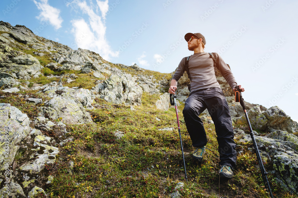 A bearded man in sunglasses and a cap with a backpack stands on top of a rock and looks into a rocky valley high in the mountains. The concept of tourism and easy trekking in the mountains outdoor A bearded man in sunglasses and a cap with a backpack stands on top of a rock and looks into a rocky valley high in the mountains. The concept of tourism and easy trekking in the mountains outdoor