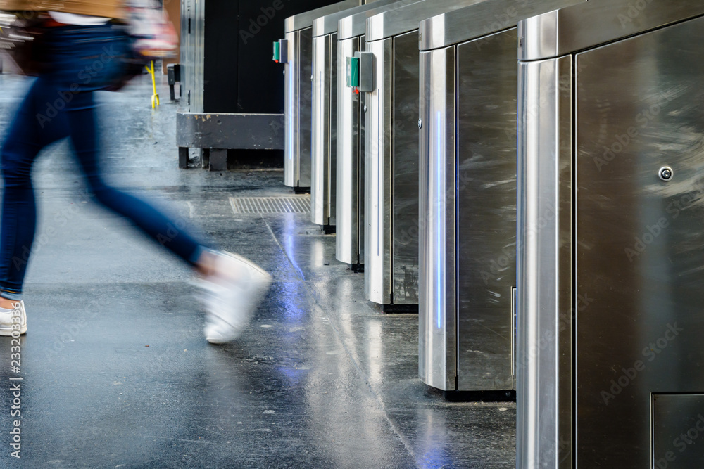 Φωτογραφία Side view of a young woman in blue jeans and white sneakers passing through stainless steel ticket gates in a subway station with motion blur