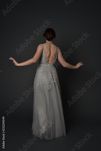 full length portrait of brunette girl wearing beautiful long gown, standing pose with background to the camera on grey studio background.