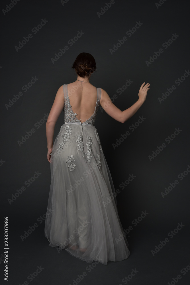 Naklejka premium full length portrait of brunette girl wearing beautiful long gown, standing pose with background to the camera on grey studio background.