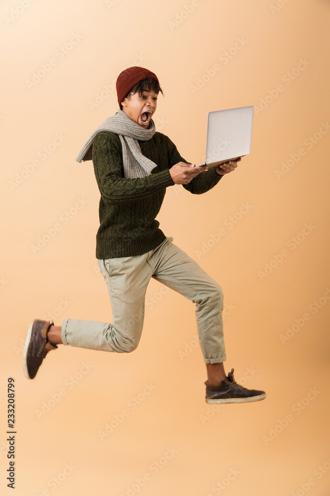 Full length photo of excited african american guy wearing hat and scarf walking with laptop, isolated over beige background Full length photo of excited african american guy wearing hat and scarf walking with laptop, isolated over beige background
