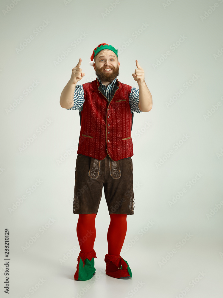 Fototapeta premium The happy smiling friendly man dressed like a funny gnome or elf pointing up on an isolated gray studio background. The winter, holiday, christmas concept