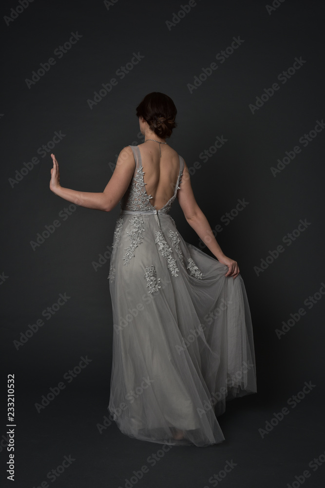 full length portrait of brunette girl wearing beautiful long gown, standing pose with background to the camera on grey studio background. full length portrait of brunette girl wearing beautiful long gown, standing pose with background to the camera on grey studio background.