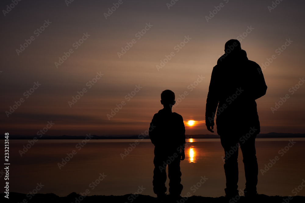 Silhouettes of the father and his little son stand on the bank of the river and watch the beautiful sunset.