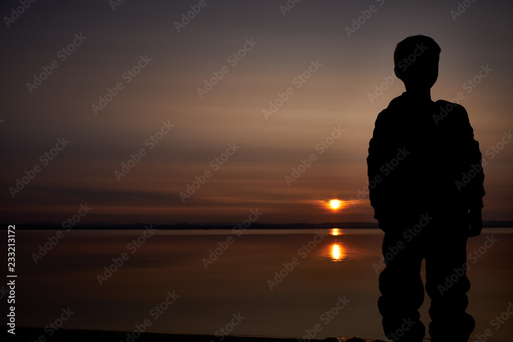  Silhouette of little boy stands in the shore of sea and looks on the sunset. Outdoors. Lifestyles concept.