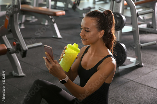 Portrait of athletic woman with protein shake and smartphone in gym