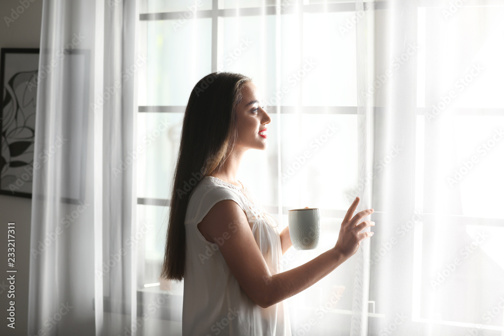Young woman standing near window with curtains at home