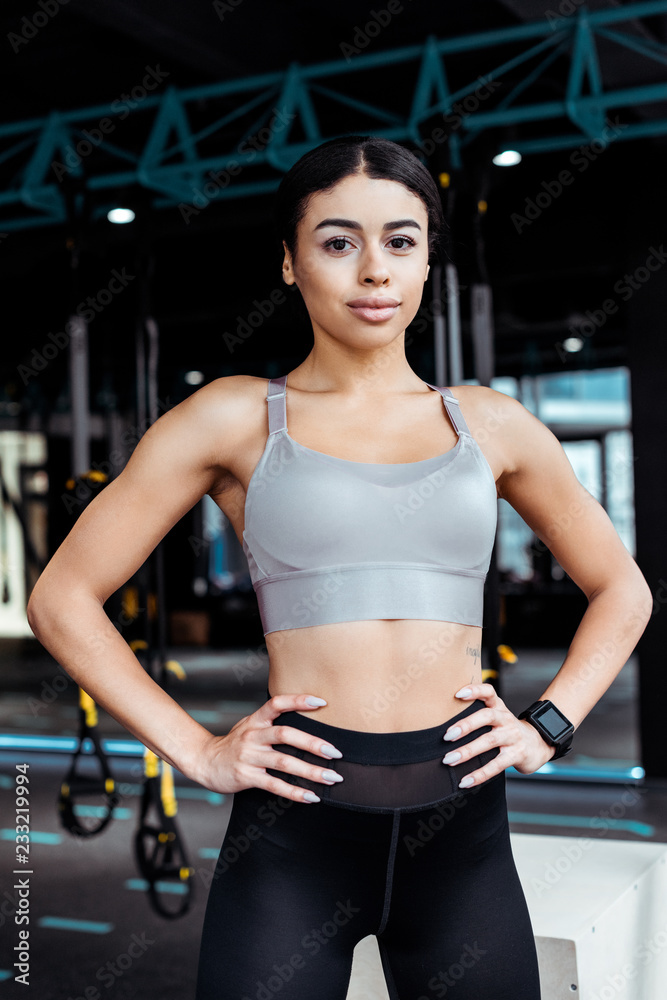 Attractive sportive girl standing akimbo while looking at camera in fitness gym