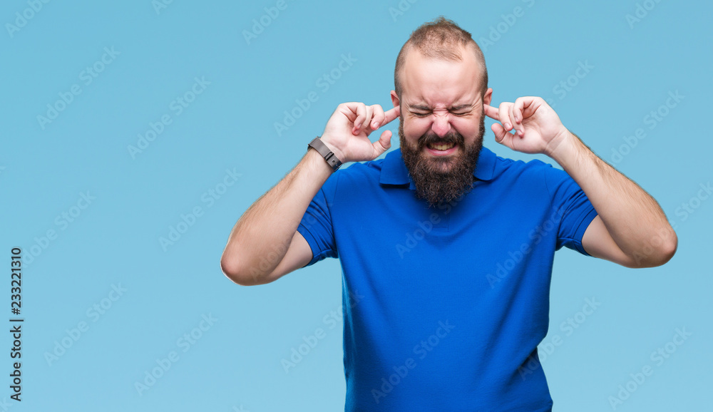 Young caucasian hipster man wearing blue shirt over isolated background covering ears with fingers with annoyed expression for the noise of loud music. Deaf concept. Young caucasian hipster man wearing blue shirt over isolated background covering ears with fingers with annoyed expression for the noise of loud music. Deaf concept.