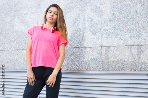 Tired jogger having break. Beautiful young Latin girl with headphones around neck looking at camera, street wall in background. Sport and active lifestyle concept