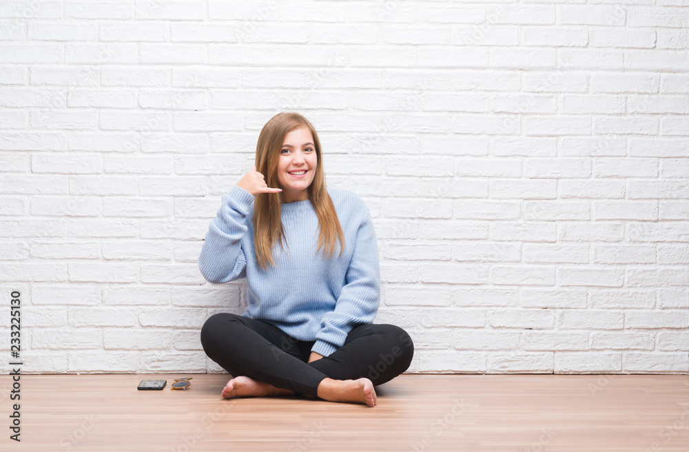 Young adult woman sitting on the floor in autumn over white brick wall smiling doing phone gesture with hand and fingers like talking on the telephone. Communicating concepts. Young adult woman sitting on the floor in autumn over white brick wall smiling doing phone gesture with hand and fingers like talking on the telephone. Communicating concepts.