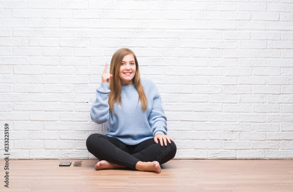 Young adult woman sitting on the floor in autumn over white brick wall pointing finger up with successful idea. Exited and happy. Number one. Young adult woman sitting on the floor in autumn over white brick wall pointing finger up with successful idea. Exited and happy. Number one.