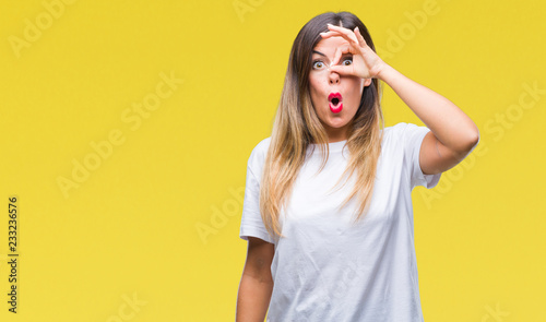 Young beautiful woman casual white t-shirt over isolated background doing ok gesture shocked with surprised face, eye looking through fingers. Unbelieving expression.