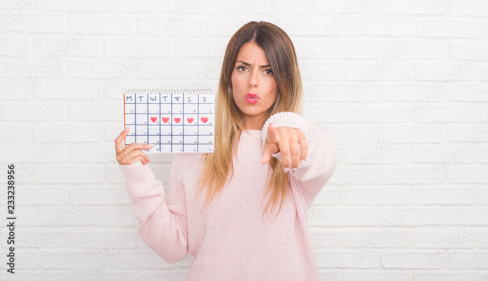 Young adult woman over white brick wall holding period calendar pointing with finger to the camera and to you, hand sign, positive and confident gesture from the front Young adult woman over white brick wall holding period calendar pointing with finger to the camera and to you, hand sign, positive and confident gesture from the front