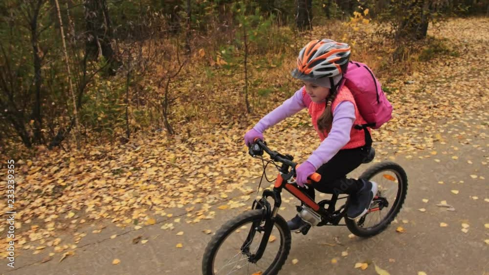 One caucasian children rides bike road in autumn park. Little girl riding black orange cycle in forest. Kid goes do bicycle sports. Biker motion ride with backpack and helmet. Mountain bike hardtail. One caucasian children rides bike road in autumn park. Little girl riding black orange cycle in forest. Kid goes do bicycle sports. Biker motion ride with backpack and helmet. Mountain bike hardtail.