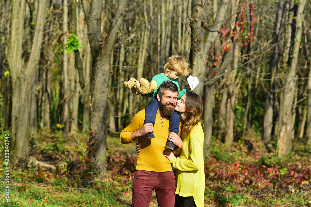 Family love concept. Mother kiss father piggybacking little child in park, family love. Soft kiss from wife Family love concept. Mother kiss father piggybacking little child in park, family love. Soft kiss from wife