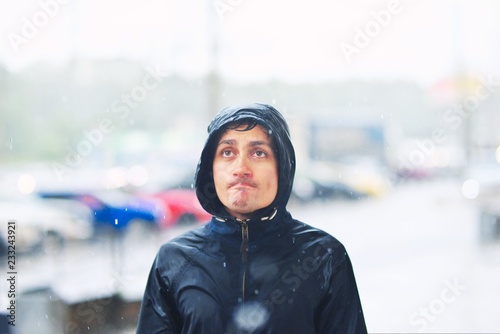 Portrait of a young man in a jacket with a hood in the rain on blurred background city street, close-up.