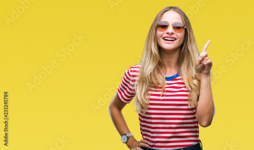 Young beautiful blonde woman wearing sunglasses over isolated background pointing finger up with successful idea. Exited and happy. Number one.