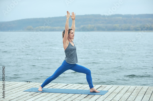 Beautiful young woman practices yoga asana Virabhadrasana 1 - warrior pose 1 on the wooden deck near the lake photo