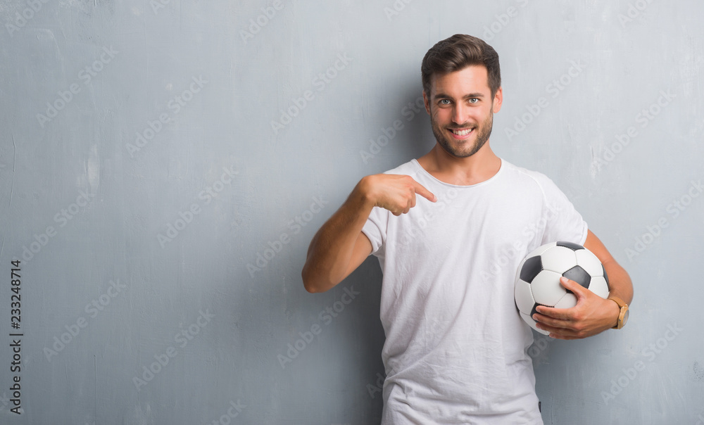 Handsome young man over grey grunge wall holding soccer football ball with surprise face pointing finger to himself