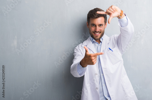 Handsome young professional man over grey grunge wall wearing white coat smiling making frame with hands and fingers with happy face. Creativity and photography concept.