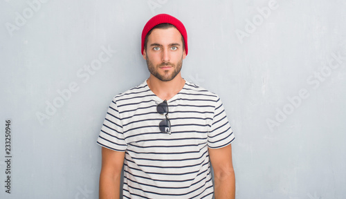 Handsome young hipster man over grey grunge wall wearing navy t-shirt and wool cap with serious expression on face. Simple and natural looking at the camera.
