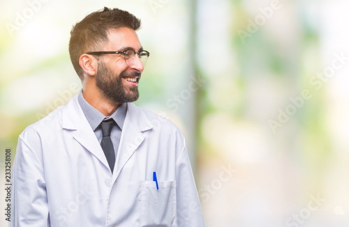 Adult hispanic scientist or doctor man wearing white coat over isolated background looking away to side with smile on face, natural expression. Laughing confident.