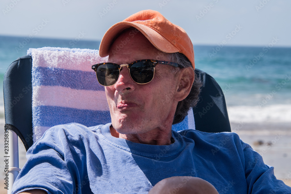 Handsome older baby boomer sad man wearing an orange cap sitting on beach chair by the ocean with a pout on his face.