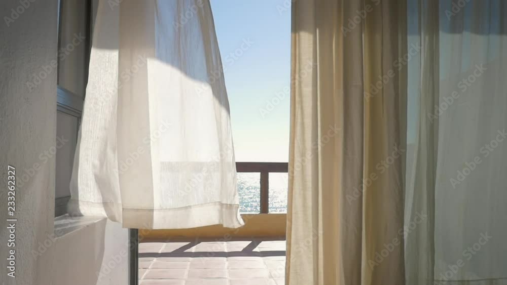 White curtains on the porch of a oceanfront resort blowing from a summer breeze in slow motion. Inside looking out windows framed by linen drapes onto the veranda of a luxury hotel with an ocean view.