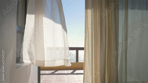 White curtains on the porch of a oceanfront resort blowing from a summer breeze in slow motion. Inside looking out windows framed by linen drapes onto the veranda of a luxury hotel with an ocean view.