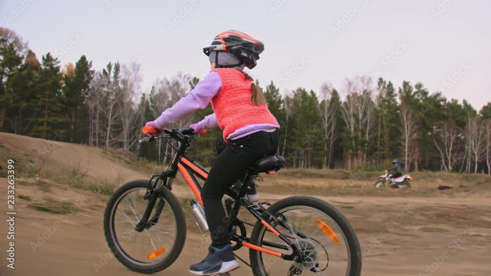 One caucasian children rides bike road track in dirt park. Girl riding black orange cycle in racetrack. Kid goes do bicycle sports. Biker motion ride with backpack and helmet. Mountain bike hardtail. One caucasian children rides bike road track in dirt park. Girl riding black orange cycle in racetrack. Kid goes do bicycle sports. Biker motion ride with backpack and helmet. Mountain bike hardtail.